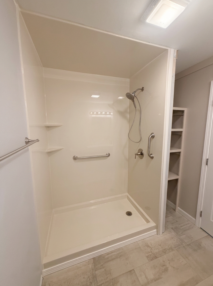 Completed accessible cream shower with seamless walls, handheld shower, grab bars, and corner shelves