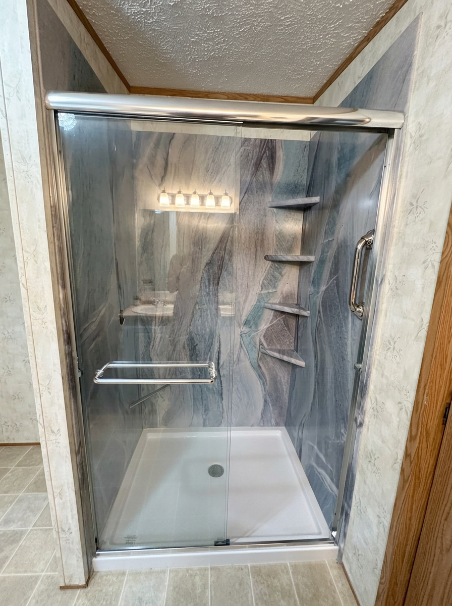 Completed blue-gray stone-look shower with chrome-framed sliding glass door, corner shelves, and white shower base